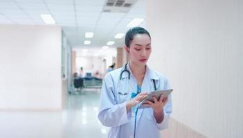 Portrait of Confident, successful professional Asian woman doctor in uniform white lab coat, stethoscope. Female physician work while stand using tablet corridor examination room in hospital medical. photo