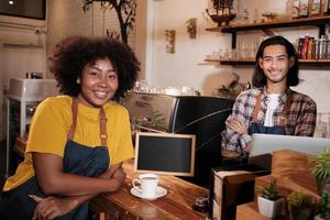 Two cafe business startup partners and friends, African American female, and Thai male baristas look at camera, arms crossed, and cheerful smiles at coffee shop counter bar, happy service staff job. photo