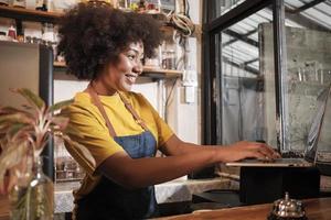 African American female cafe barista in casual apron happy with online customer order in laptop, cheerful works with smile at counter bar in coffee shop, small business startup entrepreneur lifestyle. photo