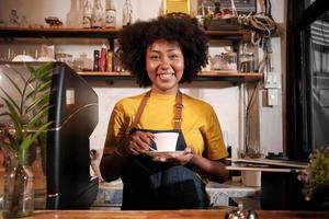 African American female barista in looks at camera, offers cup of coffee to customer with cheerful smile, happy service works in casual restaurant cafe, young small business startup entrepreneur. photo