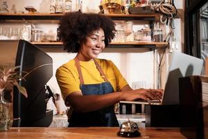 African American female cafe barista in casual apron happy with online customer order in laptop, cheerful works with smile at counter bar in coffee shop, small business startup entrepreneur lifestyle. photo