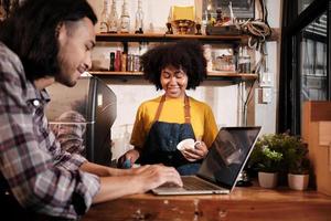 Two cafe business startup partners and friends, African American female, and Thai male baristas talk and cheerful smile together at counter bar of coffee shop, happy service job, and SME entrepreneur. photo