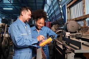 Ingenieros afroamericanos asiáticos y afroamericanos con uniforme de seguridad trabajan inspeccionando la corriente de voltaje de las máquinas, comprobando y manteniendo en la fábrica de fabricación, ocupaciones de servicio del sistema eléctrico. foto