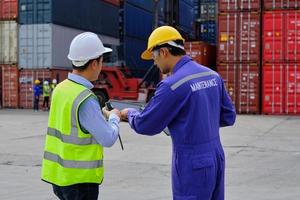 Two professional Asian male workers in safety uniforms and hard hats work at a logistics terminal with many stacks of containers, loading control shipping goods for the cargo transportation industry. photo