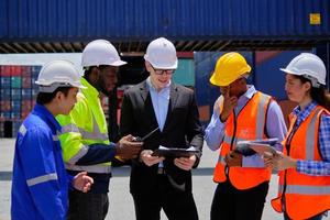 Group of multiracial workers in safety uniforms and hardhats work at logistics terminal dock with stacks of containers, loading control, and management shipping goods, cargo transportation industry. photo