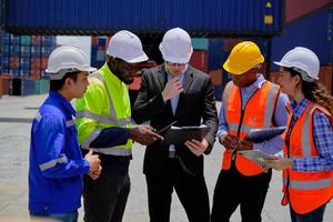 Group of multiracial workers in safety uniforms and hardhats work at logistics terminal dock with stacks of containers, loading control, and management shipping goods, cargo transportation industry. photo