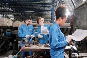 Two professional industry workers in safety uniforms and engineers' partners discussed mechanical drawing for lathe machines in manufacturing factory, with African American colleague in front of them. photo