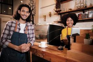 Two cafe business startup partners and friends, African American female, and Thai male baristas look at camera and cheerful smile at coffee shop counter bar as happy service job entrepreneurs. photo