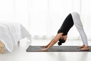 Asian women doing yoga exercise at home, stretching in Downward Facing Dog pose or adho mukha svanasana in white bedroom.  Working out wearing black shirt and white pants. Healthcare concept. photo