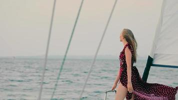 Young pretty girl on a yacht in the evening at sunset. A yacht trip on the high seas. Happy young woman smiles and raises her hand up the hill. Strong wind lifts the girl's dress. video
