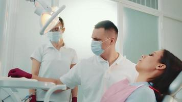 Side view of dentist and assistant in uniform treating teeth of female patient with mirror working in dental clinic. Doctor holding dental mirror, curing teeth of pretty woman. Concept of dentistry video