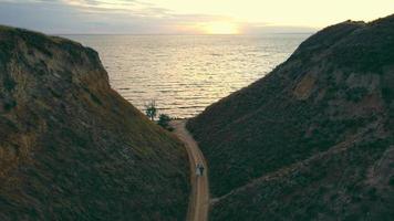 Above view of just married couple walking road between hills up to sea at sunset. Back view of man and woman in white dress approaching ocean on way through hills at twilight. Concept of destination video