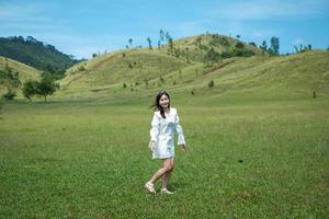 hermosa chica parada y actuando frente a la montaña calva o phu khao ya con campo de hierba verde y cielo azul. una de las atracciones turísticas naturales en la provincia de ranong, tailandia foto