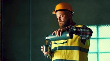 A bearded worker in a yellow helmet is resting at lunchtime and drinking tea from a thermos. 4K