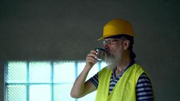 A gray-haired and bearded worker in glasses and a yellow helmet is resting at lunchtime and drinking tea from a thermos. 4K video
