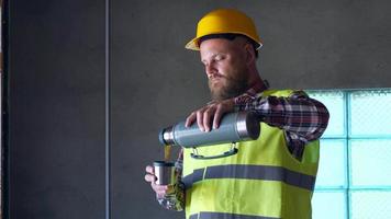 un lavoratore barbuto con un casco giallo riposa all'ora di pranzo e beve il tè da un thermos. 4k video