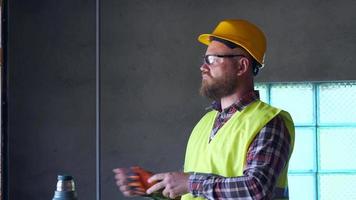 A bearded worker in a yellow helmet is resting at lunchtime and drinking tea from a thermos. He takes off his gloves and helmet and goggles. 4K video