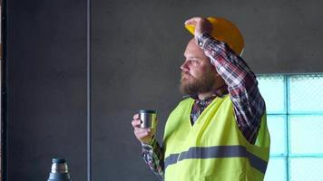 Ein bärtiger Arbeiter mit gelbem Helm ruht sich in der Mittagspause aus und trinkt Tee aus einer Thermoskanne. er zieht Handschuhe, Helm und Schutzbrille aus. 4k video