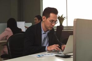 Businessman working on laptop computer sitting at office desk in her workstation with colleagues in background photo