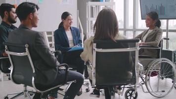 Confident business woman in wheelchair presenting during business meeting with colleagues. Asian business team celebrating success together in boardroom.
