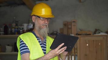 trabalhador portuário com barba em um capacete amarelo fica com um tablet pc. o capataz inspeciona. 4k video