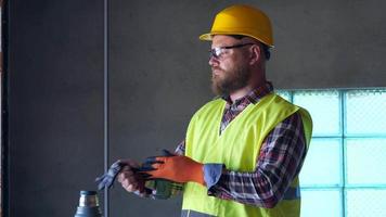 A bearded worker in a yellow helmet is resting at lunchtime and drinking tea from a thermos. He takes off his gloves and helmet and goggles. 4K video