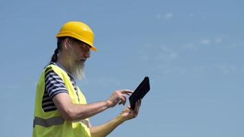 Hafenarbeiter mit Bart in einem gelben Helm steht mit einem Tablet-PC. der Vorarbeiter kontrolliert. 4k video