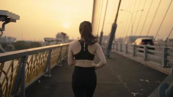 mujer asiática con un sujetador deportivo negro y pantalones negros. trotar en el puente en el centro de la ciudad por la mañana o por la noche hermosa luz del sol video