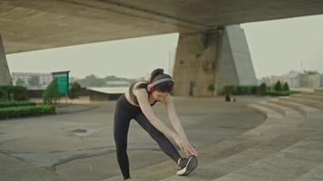 Schlanke asiatische Sportlerin, die schwarze Sportbekleidung trägt, Übungskopfhörer trägt und die Beine in einem Park in der Nähe einer Brücke über ein Flussstadtleben streckt