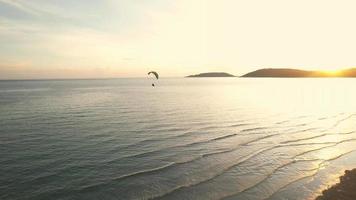 um drone voa atrás de um parapente ao longo de uma praia ao pôr do sol. vista aérea do voo de paramotor ao pôr do sol. um parapente sobrevoa uma pitoresca praia de areia ao pôr do sol. video