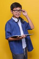 primer plano de un niño inteligente con ropa informal, auriculares y anteojos sosteniendo un libro y sonriendo a la cámara con fondo amarillo. foto