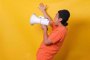 Asian casual guy isolated on yellow background shouting through a megaphone. Side view. photo