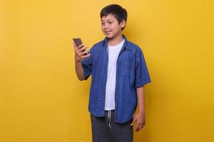 Asian boy in denim casual style is smiling while holding smartphone isolated on yellow background. Online learning concept. photo