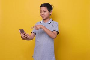 Asian boy in casual style is smiling while holding smartphone isolated on yellow background. Online learning concept. photo