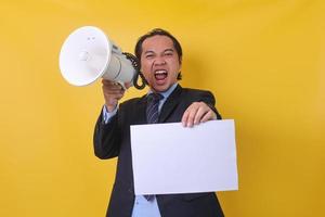 Asian businessman is yelling and shouting using megaphone and holding blank sheet paper  isolated on yellow background. Copy space and mock up. photo