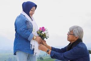 Romantic elderly couple. Husband is bend a knee and giving flower to wife. Wedding anniversary concept. photo