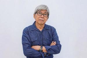 Asian senior grey-haired man in denim casual style with arms crossed  looks directly at camera with confident expression wears transparent eyeglasses isolated on white background. photo