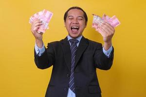 Asian businessman in suit standing against yellow background, celebrating success and holding money one hundred thousand rupiah on hands. Financial and savings concept. photo