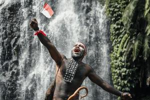 Papua man of Dani tribe say merdeka, celebrating Indonesia independence day against waterfall background. photo