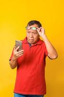 Portrait of confused Asian old man looking on smartphone while putting on glasses isolated on yellow background in casual style. photo