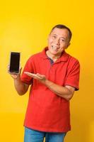 Photo of Asian old man smiling and holding smartphone with direct finger pointing to screen wearing red t-shirt isolated on yellow color background.
