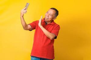 Happy Asian senior man waving hands making online video call on cell phone. Mature retired 60s  enjoying virtual meeting video call, chat, talking, looking at smartphone on yellow background. photo