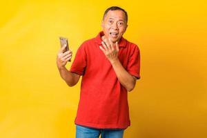 Photo of Asian grandpa hold telephone and open mouth surprised wear red t shirt isolated yellow color background.