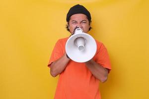 Close-up of Asian casual guy isolated on yellow background shouting through a megaphone. Front view. photo