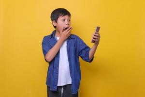 un chico asiático con un estilo vaquero informal sostiene un teléfono y muestra una expresión sorprendida aislado en un fondo amarillo foto