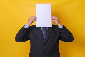 Young businessman in suit  hiding his face under blank sheet paper, isolated yellow background. Mock up and copy space. photo