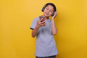 Asian boy in casual style using wireless headset and smartphone listening to music with closed eyes, yellow studio background. photo