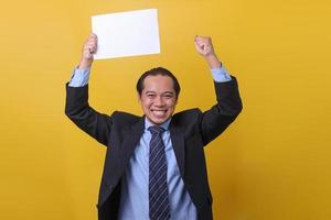Attractive Asian businessman celebrating success and holding blank white paper above the head,  yes man, success team concept after sign contract, isolated on yellow background. photo