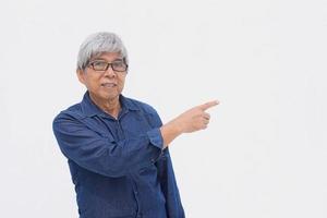 Asian senior man in denim casual style is smiling to the camera while pointing index finger isolated on white background with copy space. photo