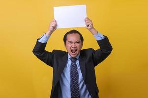 Angry and stressed businessman in suit is shouting while holding blank white paper above head,  isolated on yellow background. Failure concept. Copy space, emotional face expression. photo
